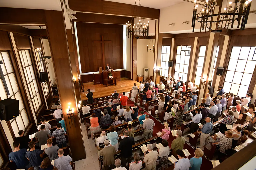 Viewed from the balcony the image looks down on a Christian congregation listens to a minister from the pulpit standing below a large wooden cross
